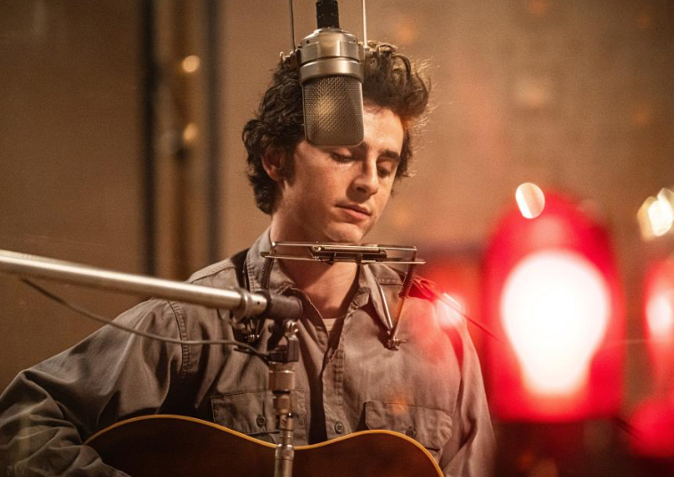 Timothée Chalamet as Bob Dylan, dressed in a 1960s-inspired outfit with a guitar and harmonica, embodying the folk-rock icon.