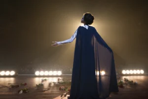 Angelina Jolie as Maria Callas standing on stage, surrounded by roses, in a moment of triumph and elegance.