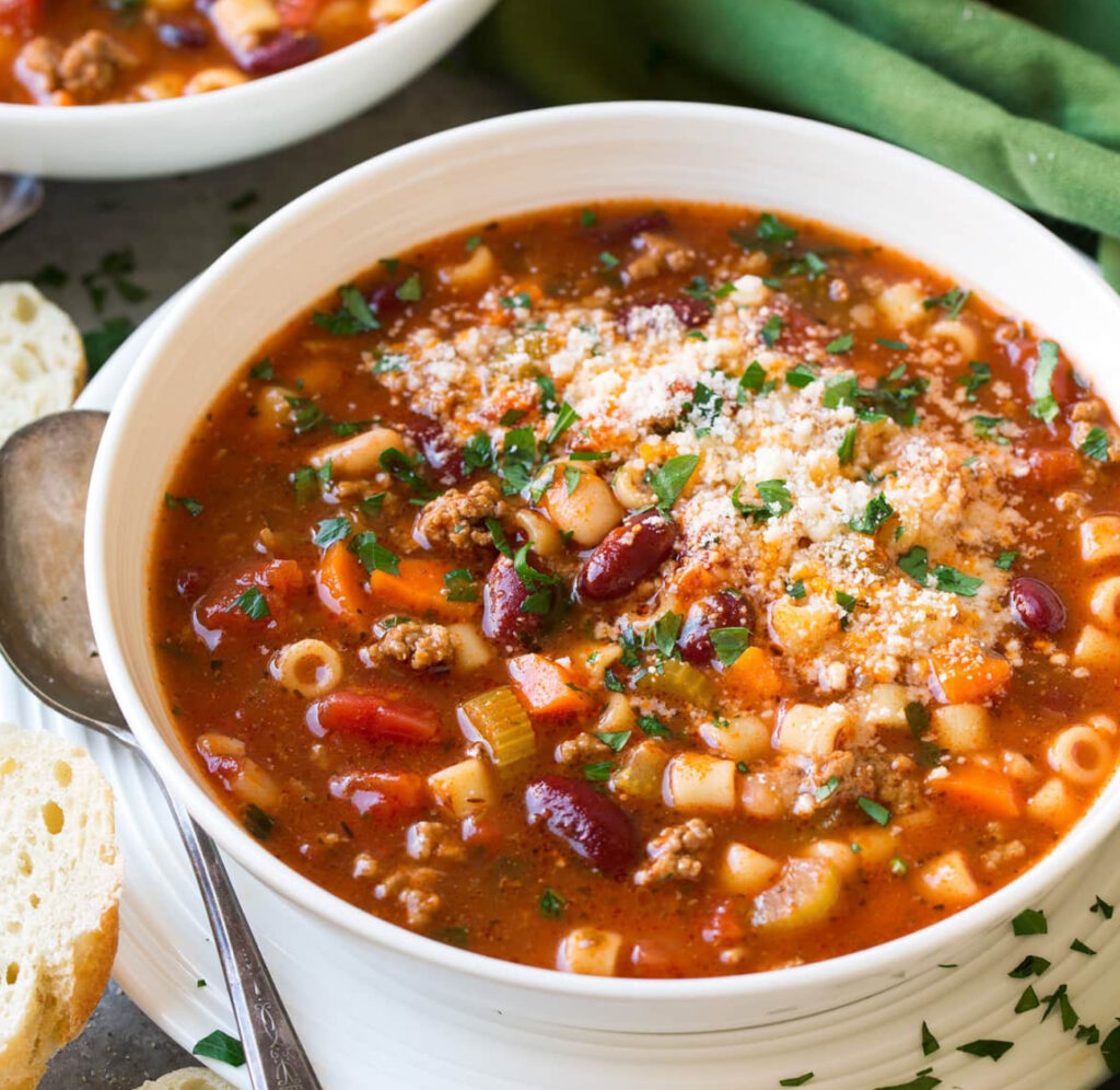 A steaming bowl of traditional Pasta Fagioli with small pasta, beans, and a rich tomato broth, garnished with parsley and Parmesan