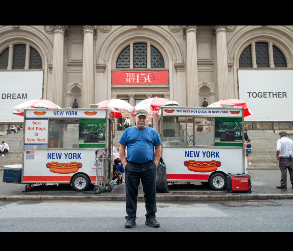 Dan Rossi, NYC’s Hot Dog King, standing beside his cart outside the Metropolitan Museum, serving tourists