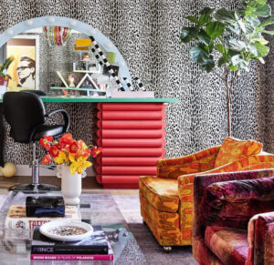 A vibrant maximalist room featuring patterned chairs, leopard-print curtains, a glass coffee table, and a colorful vanity