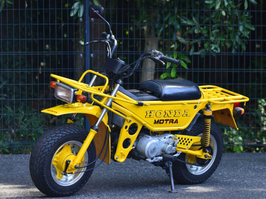 A vintage Honda CT50 Motra, featuring a rugged steel frame, front and rear cargo racks, and off-road tires in a retro setting