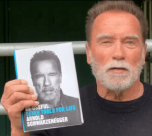 Arnold Schwarzenegger speaking on stage, emphasizing his philosophy of purpose and helping others for a meaningful life