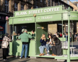 A vibrant Blank Street Coffee pop-up at a tram stop, offering commuters freshly brewed coffee and a cozy experience