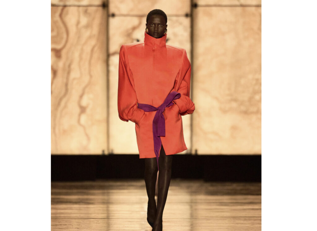 Model walks the Saint Laurent FW25 runway wearing a high-collared coat dress with soft lighting and a rock crystal backdrop