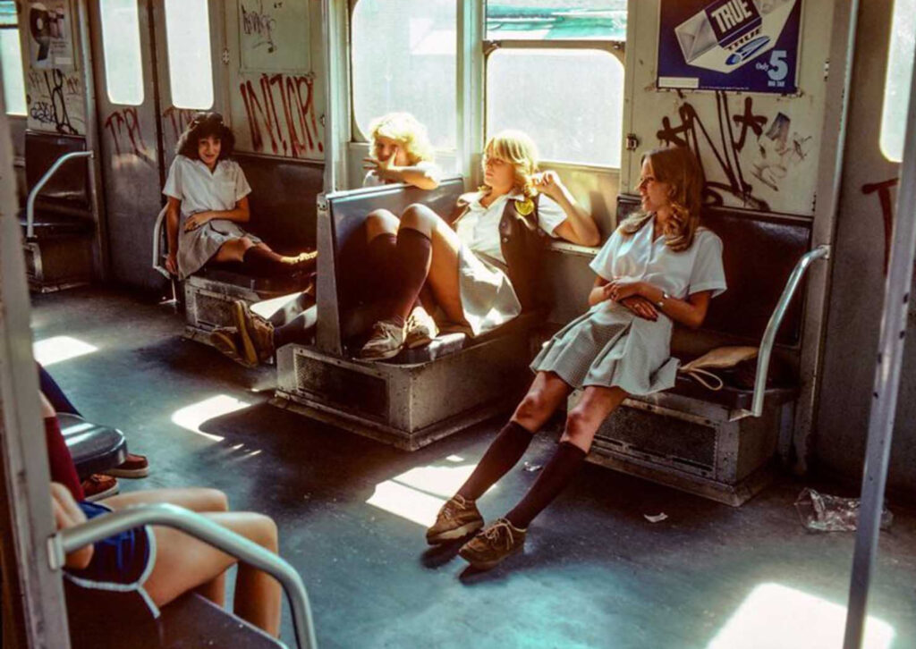 1970s NYC subway train interior with passengers seated under dim lights and graffiti covering the walls and windows