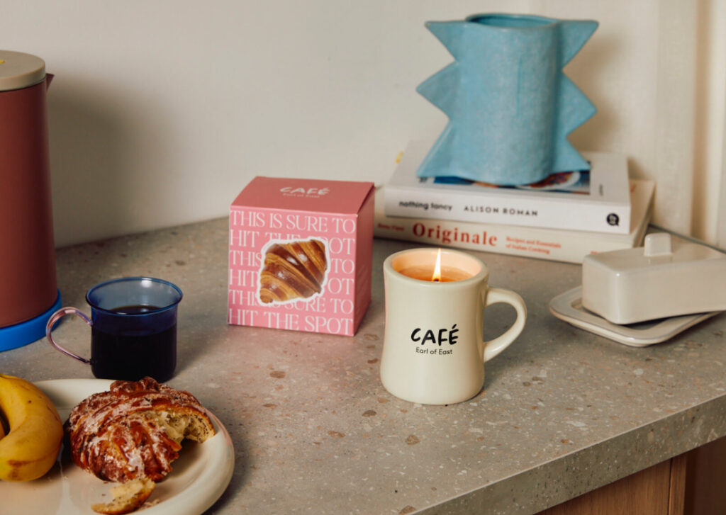 Earl of East Croissant Candle in ceramic mug with lid, placed on a wooden table beside a fresh croissant and coffee cup