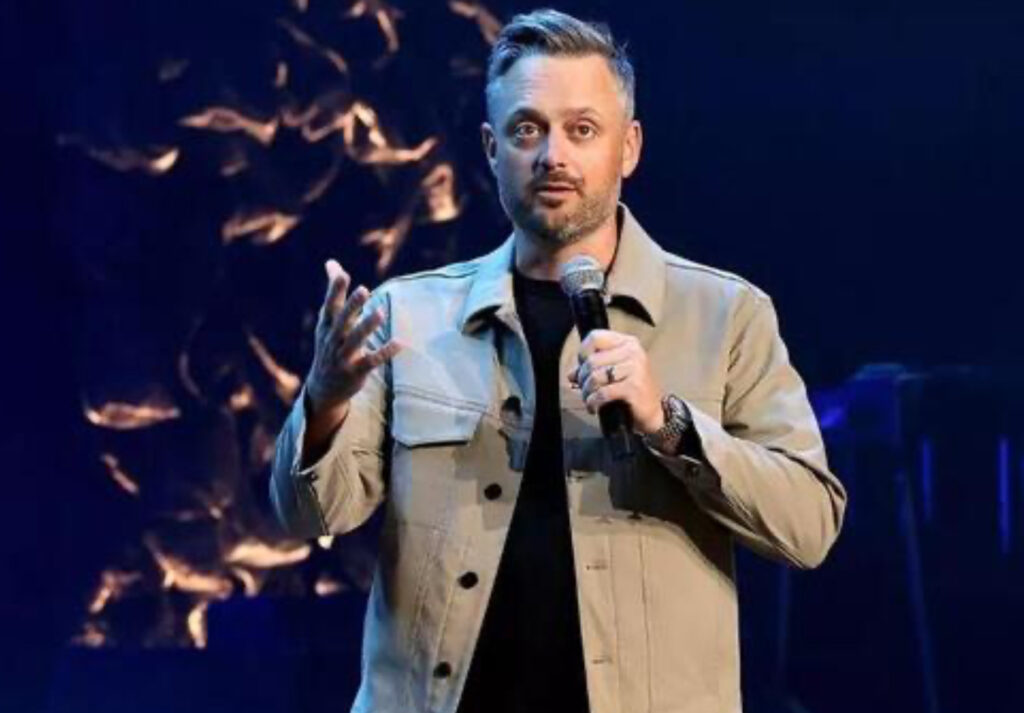 Nate Bargatze smiling onstage with a microphone, set to host the 77th Emmy Awards in Los Angeles