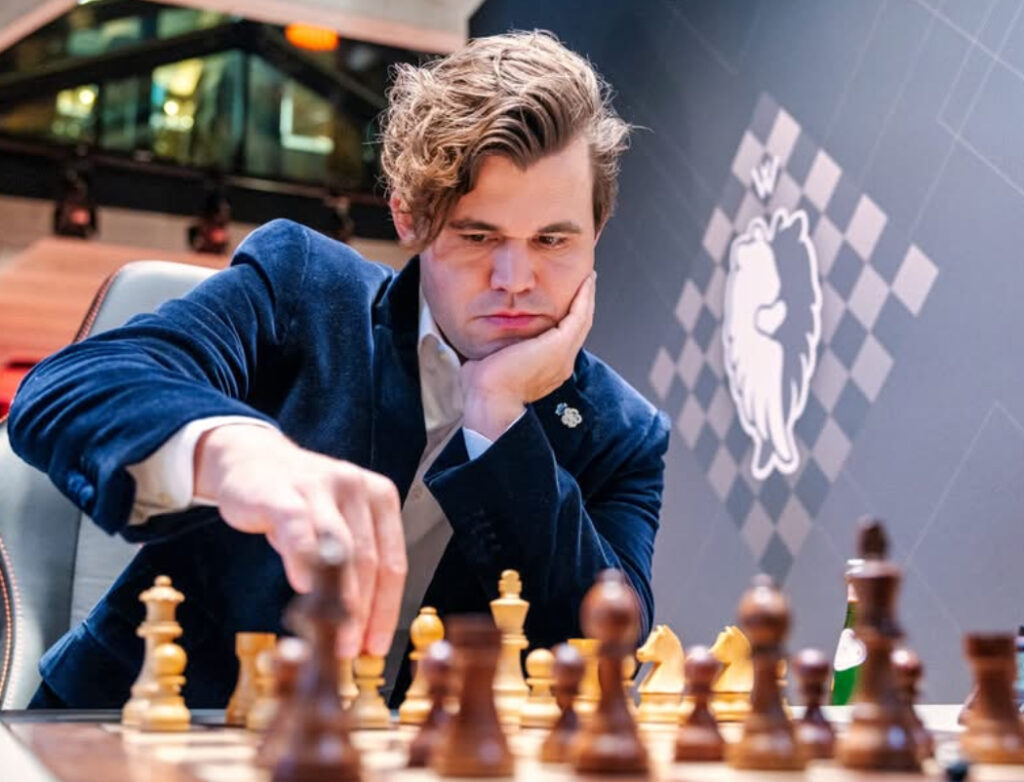 Magnus Carlsen seated at a Freestyle Chess board mid-match, smiling confidently during the G.O.A.T. Challenge event