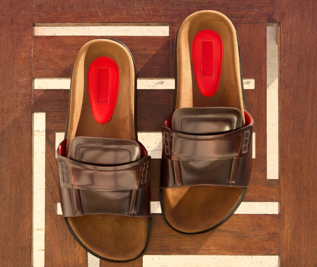 Ferrari SS25 brown leather slide sandals with red heel inserts on a wooden geometric surface, viewed from above