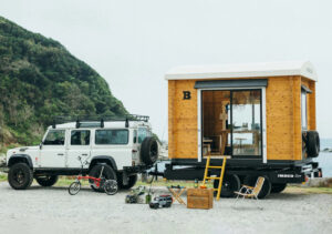 A small green RV parked in a remote wooded area, with belongings neatly arranged outside, capturing the reality of mobile living