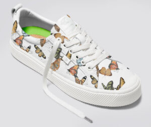 Cariuma OCA Low sneaker in white pebbled leather with butterfly print, resting against natural wood and stone backdrop