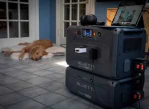 BLUETTI Apex 300 portable power station powering a fan, cooler, and mobile devices at a shaded outdoor campsite