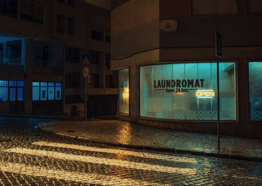 Nighttime photo of a glowing 24-hour laundromat on a cobblestone corner street with neon “OPEN” sign