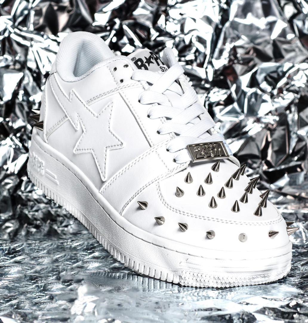 White BAPE STA™ 20th Anniversary sneaker featuring silver spikes on toe and heel, set against crinkled foil backdrop