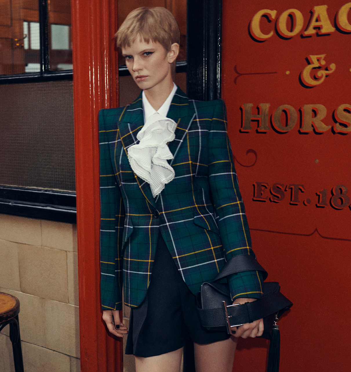 Model wearing Alexander McQueen AW25 pre-collection at The Coach & Horses pub, styled in tartan blazer and feathered trim
