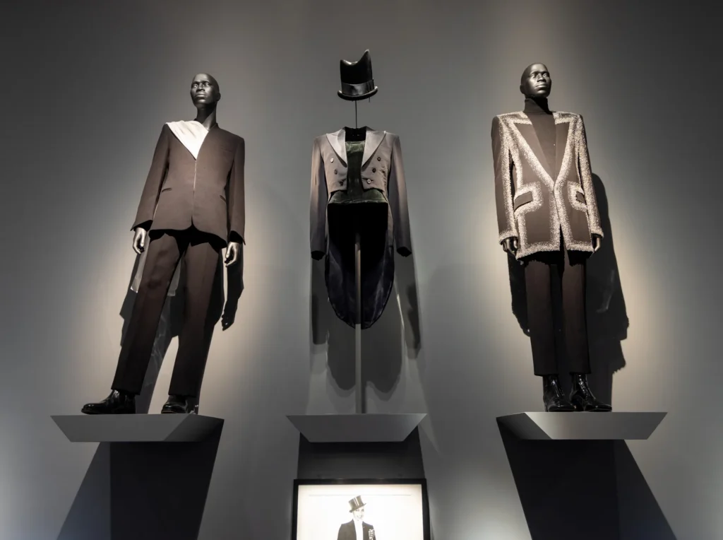 Three tailored ensembles on mannequins at the Met’s Superfine exhibition, featuring works by Maximilian Davis, Philip Treacy, and Olivier Rousteing