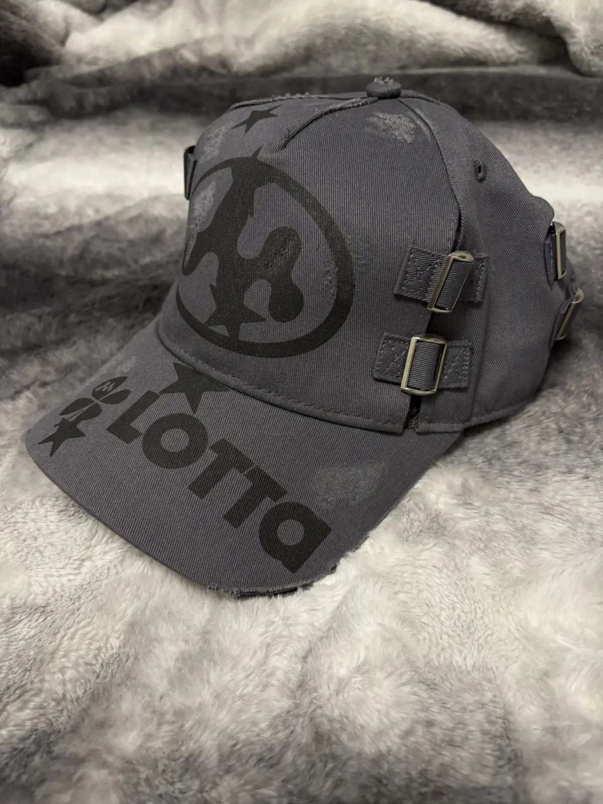 Mowalola Lotta cap in gray with abstract graphic and metal buckle details, floating on reflective surface evoking mystery