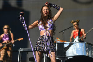 Olivia Rodrigo performing on stage in a purple checkered crop top and printed skirt, smiling and holding a lavender mic