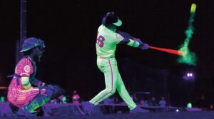 a player hitting a ball and catcher aside wearing glow-in-the-dark baseball at night under ultraviolet lighting, with neon uniforms and glowing equipment
