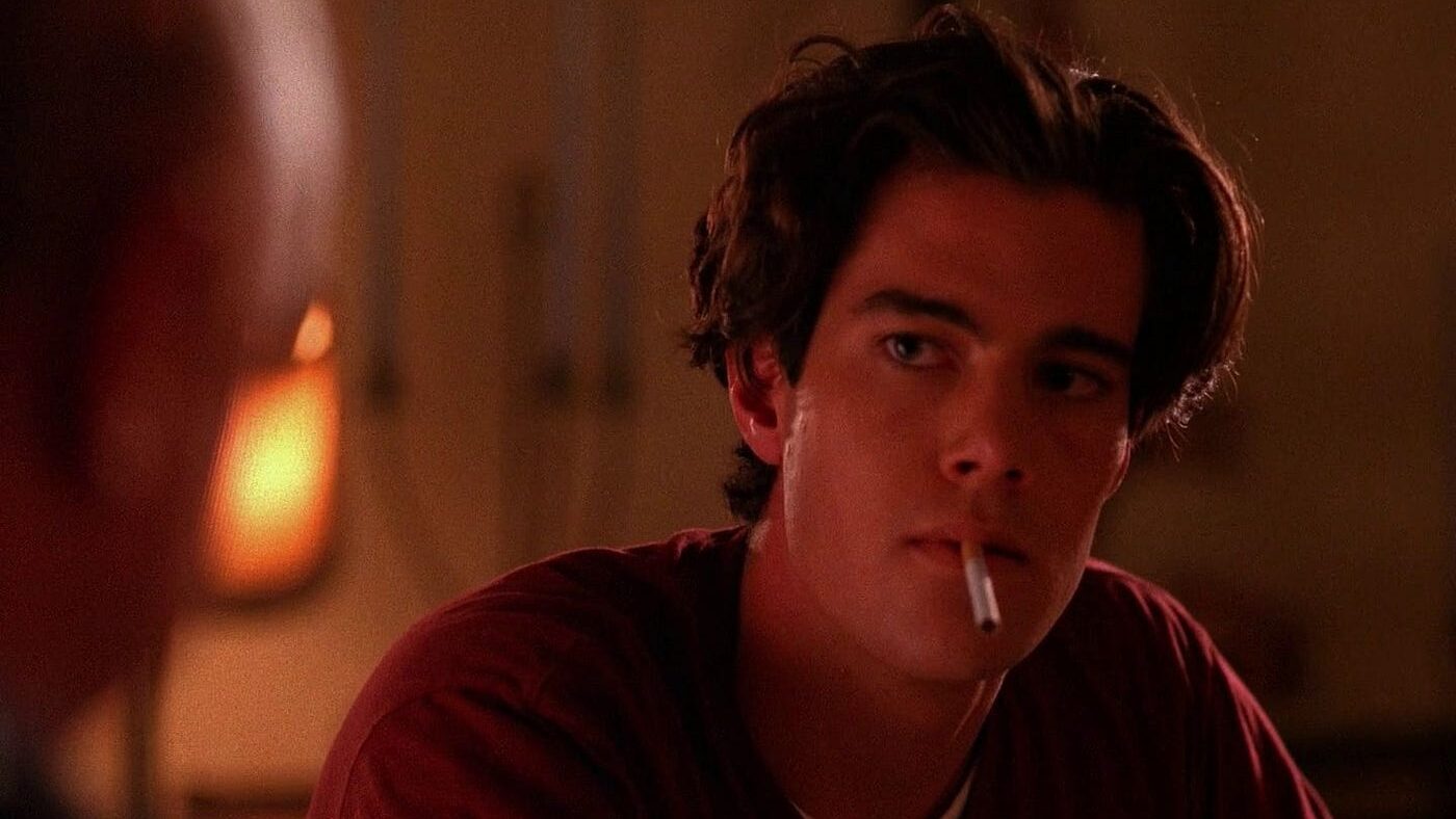 Dana Ashbrook as Bobby Briggs in Twin Peaks, smirking with a cigarette, framed in soft retro lighting with a 90s noir mood