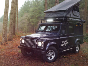 A 2025 Land Rover Defender 110 parked by a scenic overlook with rooftop camping gear and open cargo tailgate in summer light