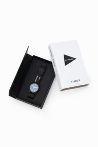 A sleek Timex × And Wander Expedition North Field Post Solar watch in matte titanium finish, featuring luminous numerals and a durable fabric strap with reflective stitching, set against a minimalist outdoor-inspired background
