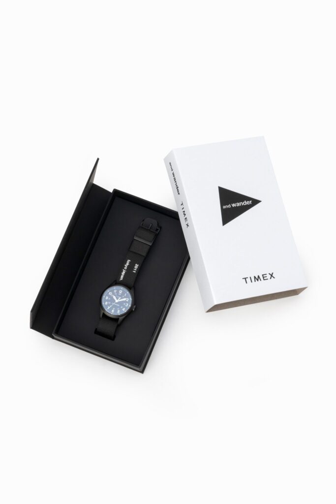A sleek Timex × And Wander Expedition North Field Post Solar watch in matte titanium finish, featuring luminous numerals and a durable fabric strap with reflective stitching, set against a minimalist outdoor-inspired background