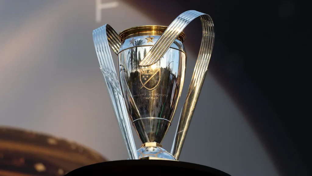 The MLS Cup trophy, polished silver with gold accents and two curved handles, standing against a soft stadium backdrop, symbolizing ultimate championship victory in Major League Soccer