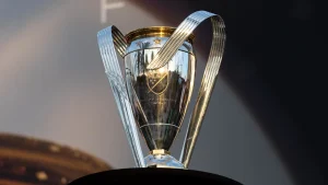 The MLS Cup trophy, polished silver with gold accents and two curved handles, standing against a soft stadium backdrop, symbolizing ultimate championship victory in Major League Soccer