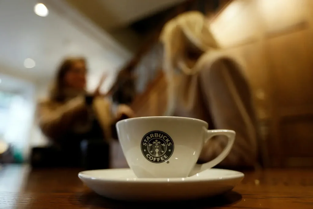 Close-up of a white Starbucks coffee cup with the logo, placed on a saucer on a wooden table, with two people blurred in the background having a conversation inside a warm, softly lit café setting