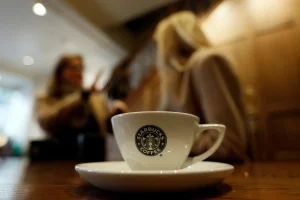 Close-up of a white Starbucks coffee cup with the logo, placed on a saucer on a wooden table, with two people blurred in the background having a conversation inside a warm, softly lit café setting