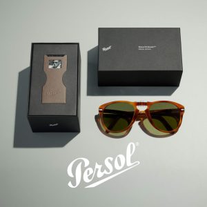 A flat lay featuring the Persol Steve McQueen™ Special Edition sunglasses with amber-colored acetate frames and green lenses, positioned beside premium black packaging boxes—one open to reveal a photo insert of Steve McQueen and embossed branding, and one closed. The white Persol logo is prominently displayed on the surface beneath