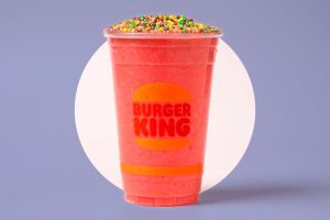 A vibrant pink frozen Burger King drink in a clear cup with the Burger King logo, topped generously with colorful Nerds candy pieces, set against a light purple background