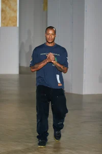 Designer Heron Preston walking on the runway wearing a navy 'Protect Arctic Wildlife' T-shirt, black cargo-style pants, and sneakers, hands clasped in front, at the close of his fashion show