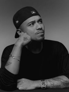 Black-and-white portrait of designer Rhuigi Villaseñor wearing a backward baseball cap and a dark shirt, resting his chin thoughtfully on his tattooed arm, showcasing his luxury wristwatch