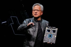 Nvidia CEO Jensen Huang wearing a black leather jacket on stage, holding a large AI processor board during a keynote presentation