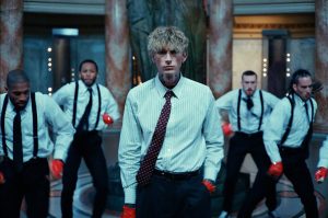 Machine Gun Kelly wearing a white striped shirt and red polka-dot tie stands in front of four backup dancers dressed in white shirts, black suspenders, and red gloves, performing in an ornate hall with marble columns and patterned floors
