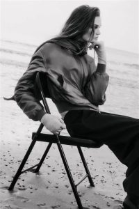 Black-and-white image of a woman seated on a beach in a scarf-detail padded silk bomber jacket, showcasing the jacket’s draped scarf and satin-like texture
