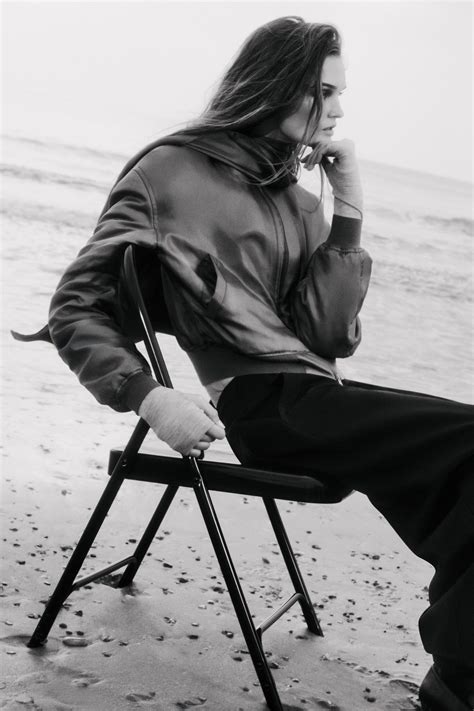 Black-and-white image of a woman seated on a beach in a scarf-detail padded silk bomber jacket, showcasing the jacket’s draped scarf and satin-like texture