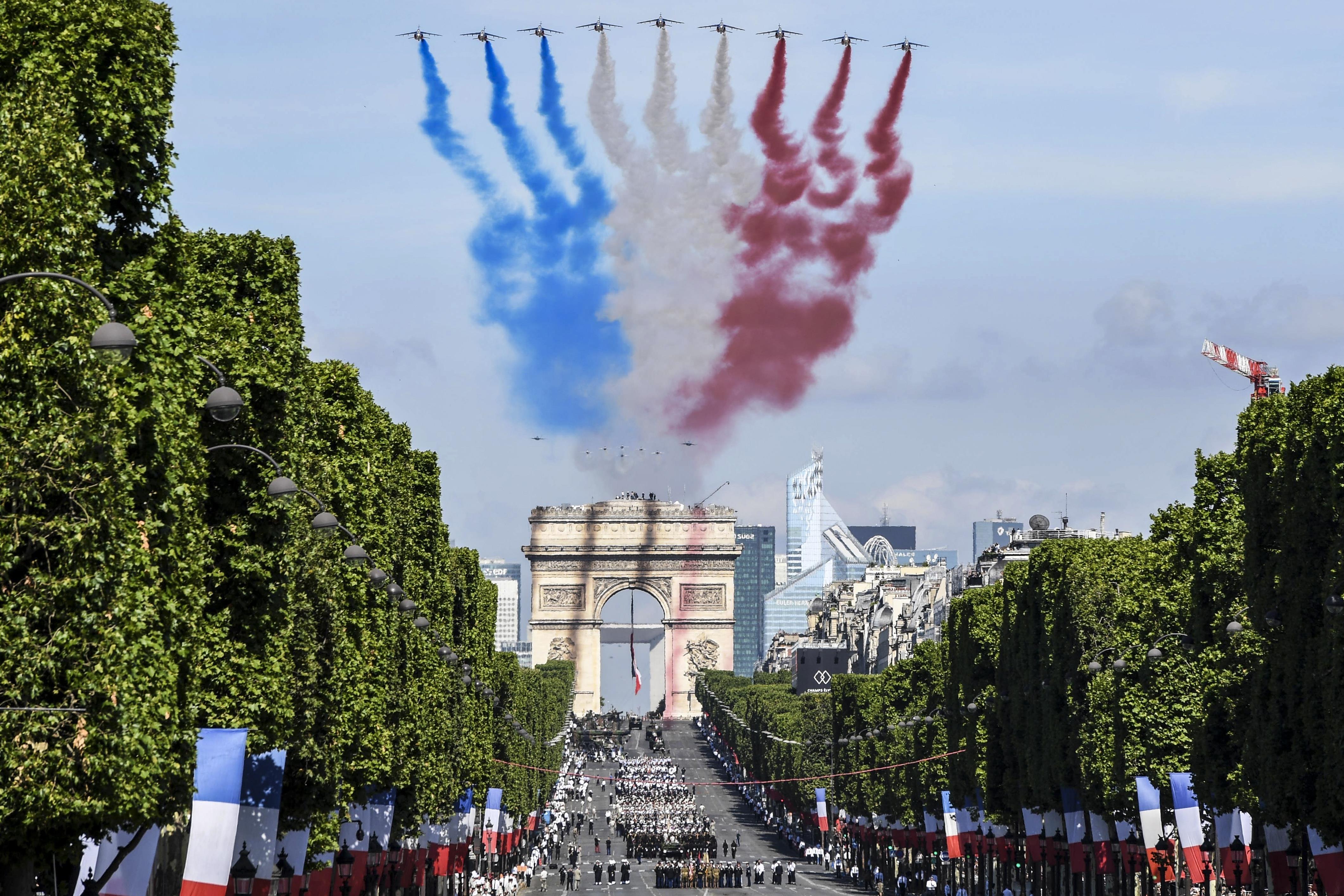 A sharp, high-resolution image of Bastille Day celebrations in Paris showing nine military jets flying over the Arc de Triomphe, leaving blue, white, and red smoke trails symbolizing the French flag, with a military parade marching down the Champs-Élysées lined with French flags and lush green trees