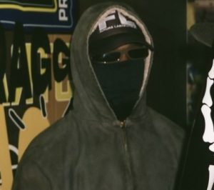 Travis Scott in hoodie, sunglasses, and face covering, standing in front of “TRAGG” backdrop