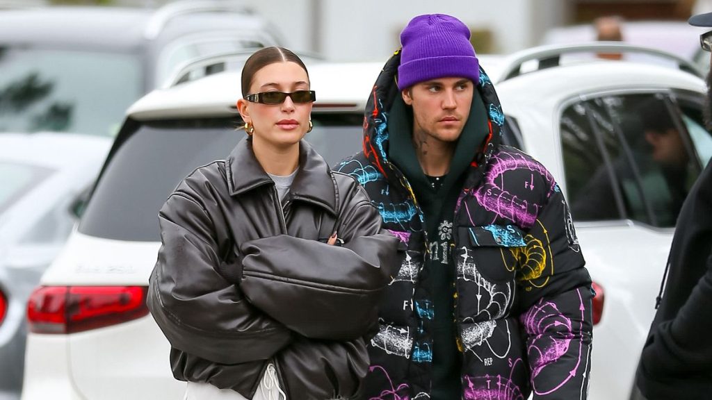 Hailey Bieber wearing a black oversized leather jacket and narrow sunglasses standing next to Justin Bieber in a colorful printed puffer jacket and purple beanie, both looking serious while walking outdoors