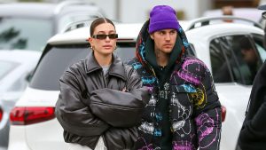 Hailey Bieber wearing a black oversized leather jacket and narrow sunglasses standing next to Justin Bieber in a colorful printed puffer jacket and purple beanie, both looking serious while walking outdoors