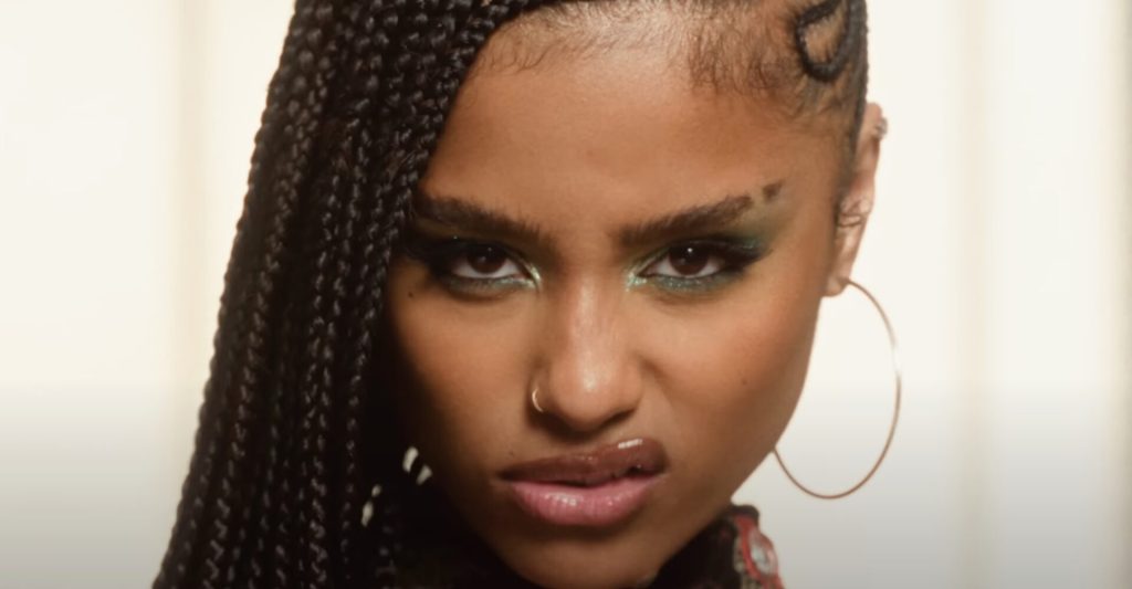 Close-up of Tyla with long braided hair, hoop earrings, and shimmering green eye makeup, staring intensely at the camera with a fierce, confident expression