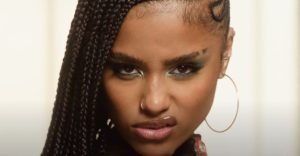 Close-up of Tyla with long braided hair, hoop earrings, and shimmering green eye makeup, staring intensely at the camera with a fierce, confident expression