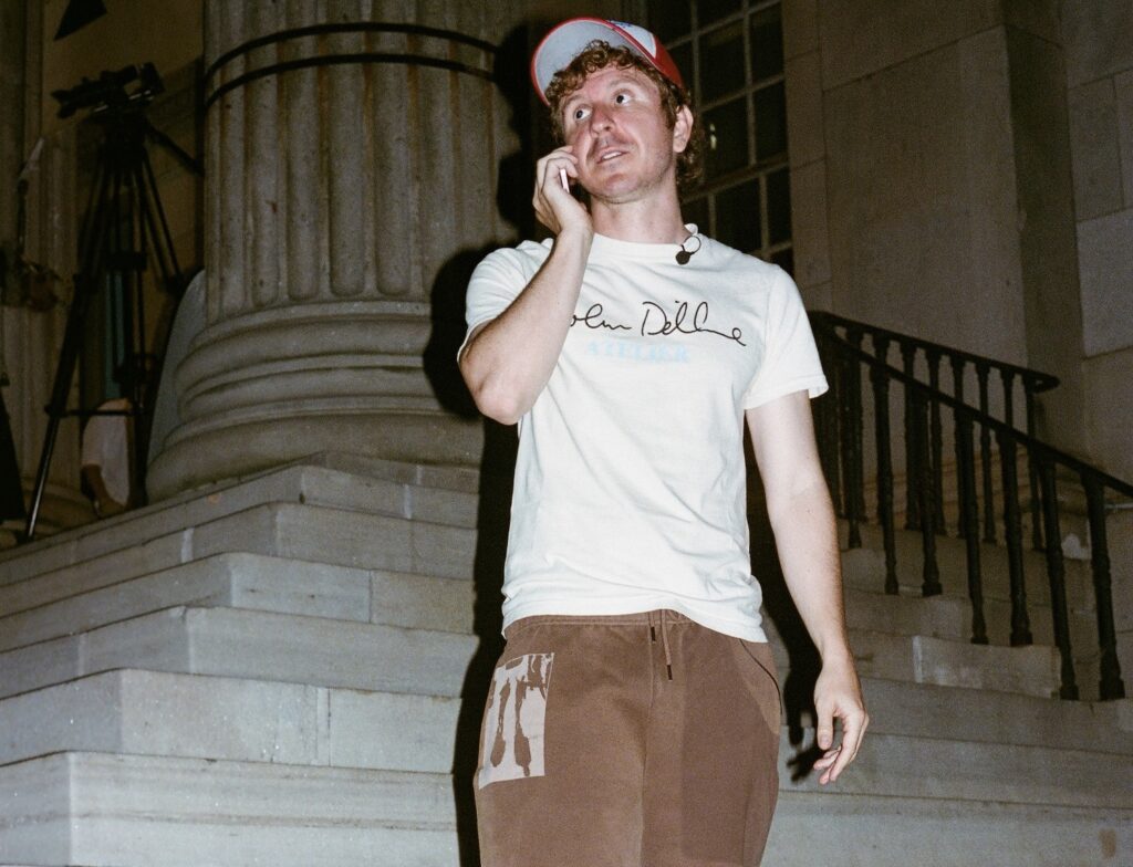 Kid Super wears a white T-shirt with “Colm Dillane” written across the chest, brown sweatpants with a graphic patch, and a red-and-blue baseball cap