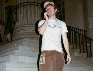 Kid Super wears a white T-shirt with “Colm Dillane” written across the chest, brown sweatpants with a graphic patch, and a red-and-blue baseball cap