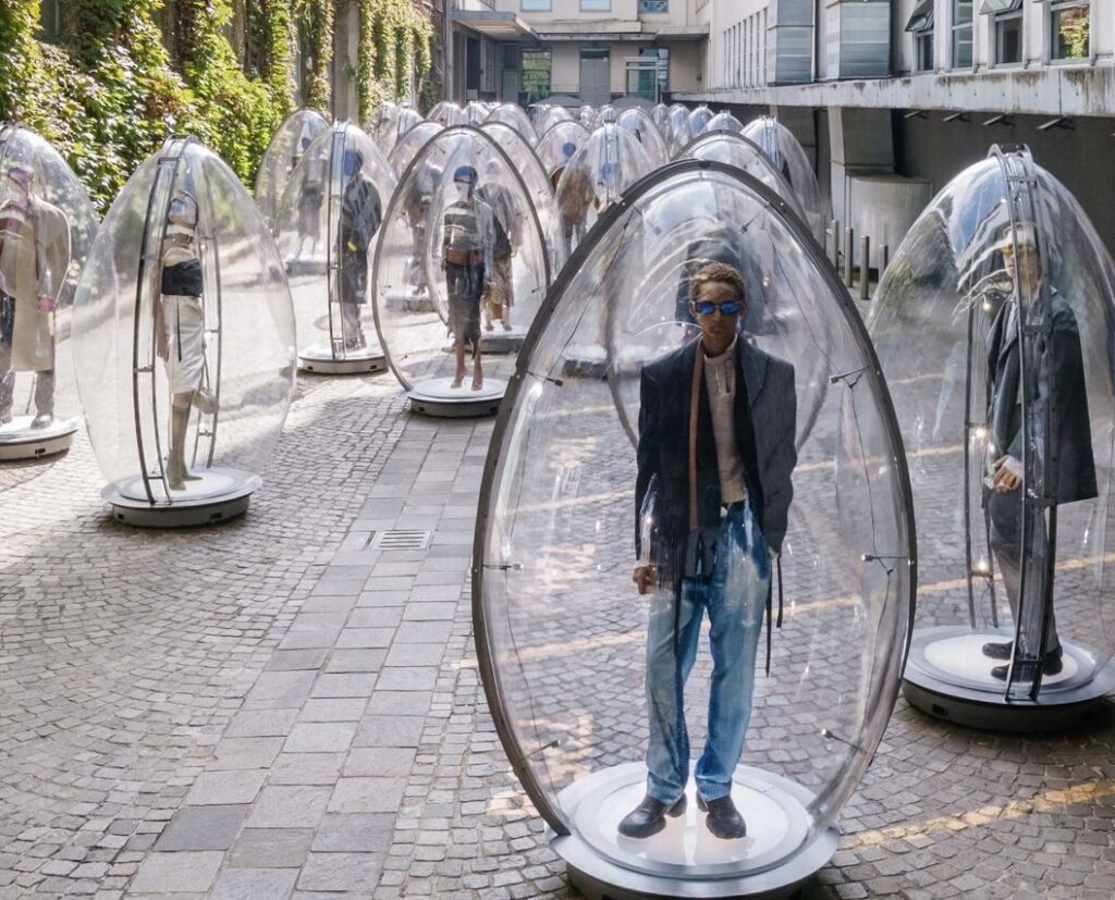 Transparent egg-shaped pods displaying Diesel SS26 runway looks in a Milan street, with models standing inside futuristic vessels.