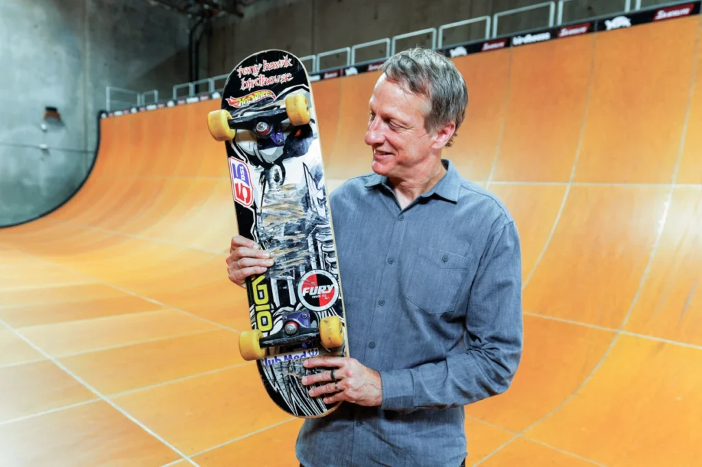 Tony Hawk smiling while holding his Birdhouse Falcon 2 skateboard, the historic deck used to land the first-ever 900 at the 1999 X Games, standing on a vert ramp.